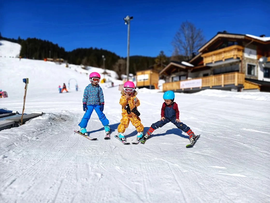 První lyžování s dětmi Tři děti v lyžařských helmách stojí na lyžích v pluhu na mírném zasněženém svahu.