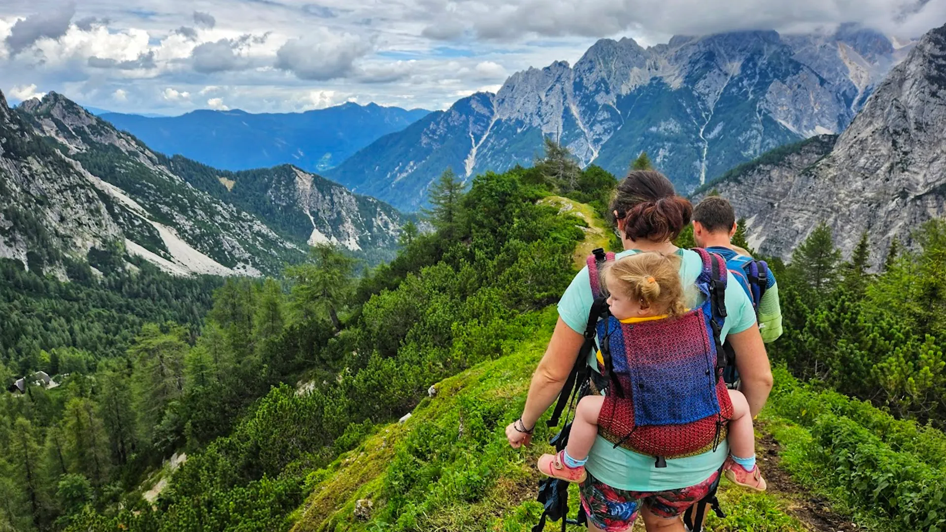 Turistika s dětmi: když přijde únava Rodič nese dítě v nosítku na horské stezce s výhledem do údolí, turistika s dětmi v horách